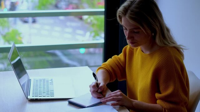 Concentrated female student taking notes in a notebook while working on her laptop computer, studying for an exam in a modern library or cafe with a city view from the window