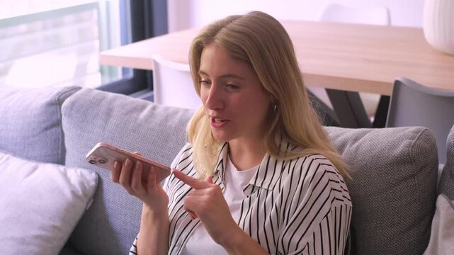 Cheerful young woman sitting on a comfortable sofa at home recording a voice message on her smartphone, smiling and gesturing while talking on speakerphone with a friend or colleague