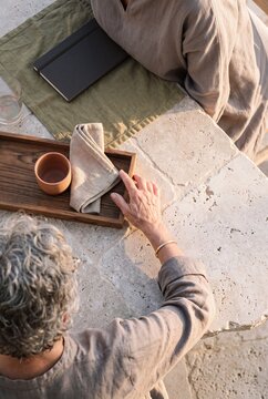 High angle view of a woman with gray hair sitting at a rustic travertine stone table with a wooden tray, terracotta cup, and notebook outdoors