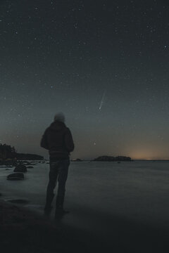 Merikula, Harju maakond, Estonia - 21 October 2025: View of a person standing on the beach at night, observing a comet in the starry sky over the sea.