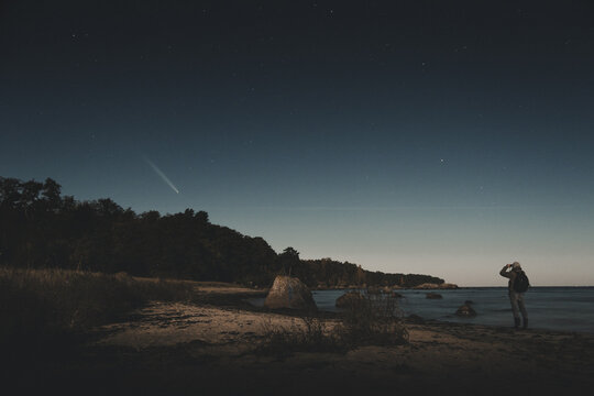 View of a person standing on the beach at night observing a comet in the starry sky above the coastline in Merikula, Harju maakond, Estonia.