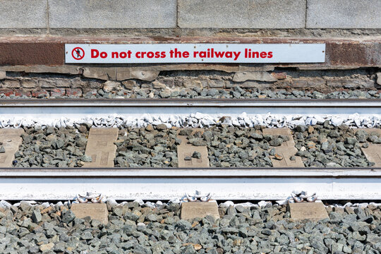 Close-up of a railway track with concrete sleepers and gravel ballast, featuring a white warning sign with red text that reads "Do not cross the railway lines" on the platform wall.