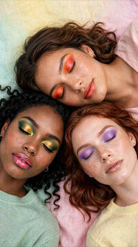 Three young diverse women with vibrant colorful eye makeup and closed eyes lying down on pastel background