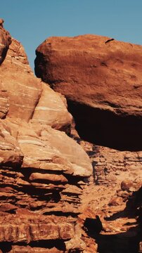 Large boulder sits on top of rocky formations in a dry desert area. Sunlight highlights the textures of the rocks. Clear blue sky is visible above the scene.