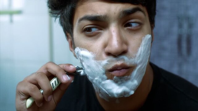 Close-up shot of a boy with shaving cream on face and using a razor - routine hygiene concept . HD stock clip of a young man standing in front of the mirror shaving his face with white foam and razor