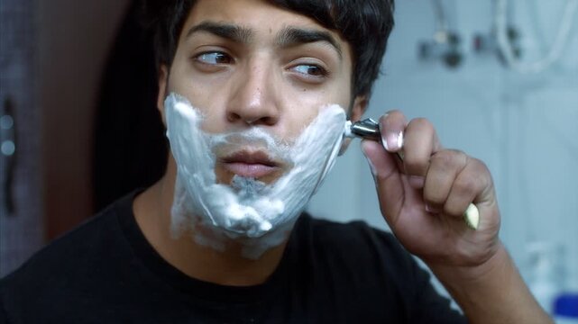 Close-up shot of a young handsome man standing in washroom and gliding razor on the face. Indian stock footage of Indian man with shaving cream on face gliding razor while looking in the mirror