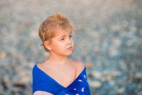 Thoughtful 5 year old boy wrapped in a blue towel on a pebble beach.