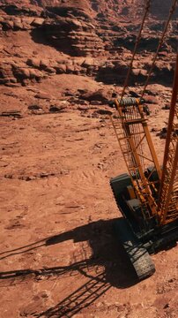 A large excavator operates on a red rocky terrain under bright sunlight. Loose soil and rocks are nearby. The scene depicts industrial work in an outdoor setting.