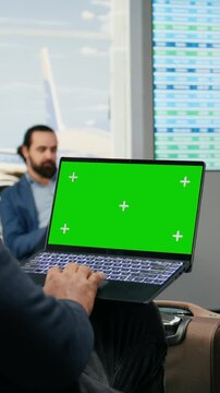 Vertical aspect: Green screen laptop appears beside a Black casual man in an airport lounge as he checks flight details and waits for an international departure while working during business travel.