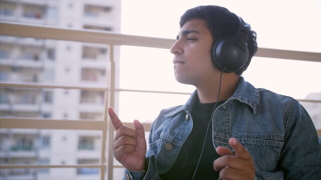 Cheerful Indian man listening to music using his headphones - technology concept. An attractive young guy in black headphones enjoying his favorite songs while sitting alone in the balcony - lifest...