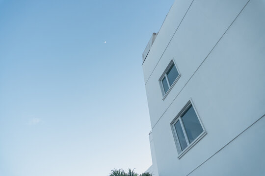 Clear sky above modern white building. Calm atmosphere with soft blue gradient. Single window reflects quiet urban stillness. Moon hangs low, adding serene contrast