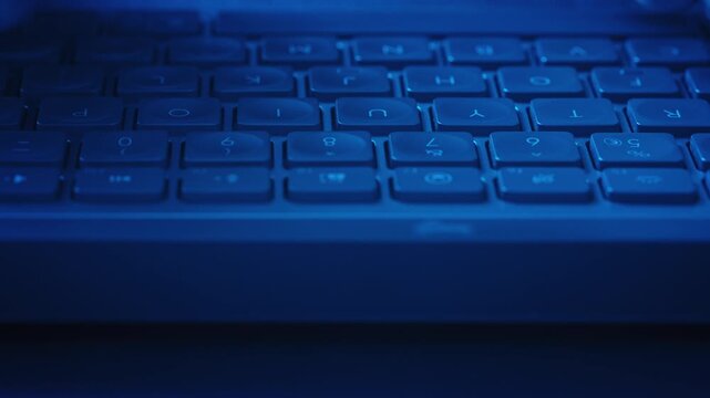 Close-up male hands typing on keyboard under blue ambient lighting while the camera slides parallel to keys. Cinematic cybersecurity, software development, technology, coding and digital workflow