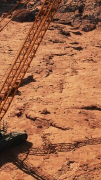 Workers operate a large crane on rocky terrain in a dry location. The ground is uneven and dusty with visible marks from the cranes movement.