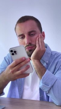 Young handsome man feeling bored and tired while scrolling through his smartphone, showing signs of digital fatigue, doomscrolling and procrastination while sitting at his desk