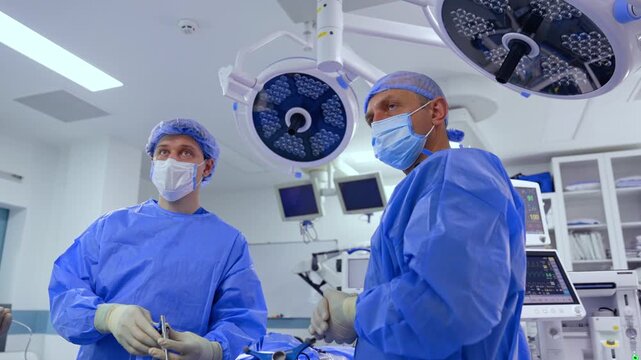 Concentrated male surgeons stand in surgical room looking at the monitor. Doctors hold the tool input into patient's body.