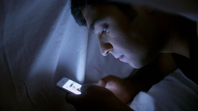 A young Indian boy / man lies under a blanket and scrolling his smartphone. 