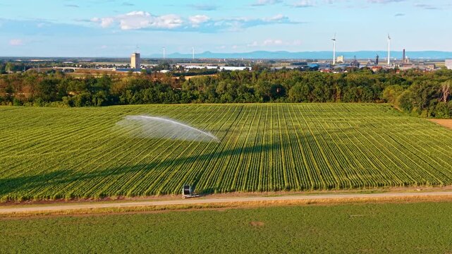 High angle view of a green field with active irrigation. A long water spray arches over agricultural crops while distant buildings and wind turbines line the horizon.