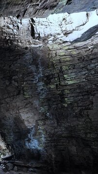 Visitors stand below a high rock ceiling in a cave, where light filters through the opening. The walls are made of layered stones, creating a rugged appearance.