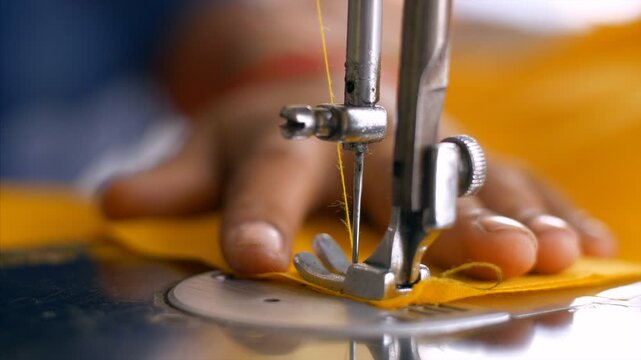 Hands of an Indian female tailor slowly working on a professional sewing machine. Closeup shot of a fashion designer carefully stitching a yellow fabric using a sewing machine in a textile workshop