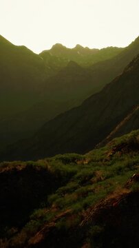 Sunset light falls on a valley surrounded by tall mountains. The landscape shows green bushes and rocky slopes. The sun casts warm colors, creating a natural scene.