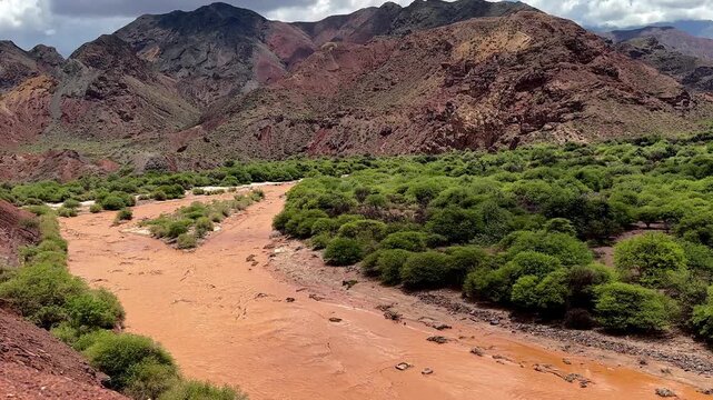 Panoramic zoom out view of a muddy river in a dry valley with green shrub and red mountains in Salta, Argentina. Scenic landscape, vivid, saturated colors. South America 