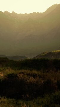 Mountains rise in the background as the sun sets, casting shadows on the landscape. Fields and grass are visible in the foreground, creating a scenic view of nature.