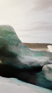Two large ice formations sit near a body of water. The sun shines down, lighting up the blue and white ice. Mountains rise in the background under a cloudy sky.