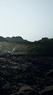A stream flows down a rocky hillside with large boulders and greenery. The sun is shining, creating a bright scene in a natural setting. The terrain is uneven and rugged.