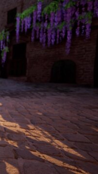 Wisteria flowers hang from a building in a village. Cobblestones form the pathway. The sun casts long shadows on the ground. The scene looks quiet and inviting.