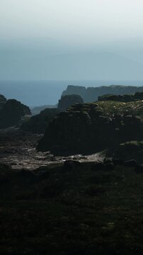 People explore rocky cliffs near the ocean on a foggy day. The coastline has a varied landscape with shadows cast by the cliffs. The waves crash against the rocks below.