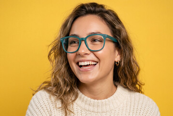 Fototapeta premium Smiling young woman wearing teal glasses and a white knitted sweater on a yellow background