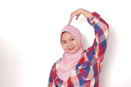 Young Asian Muslim woman wearing pink hijab smiling and making half heart shape gesture above head isolated on white background