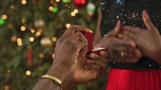 Close-up of a man presenting an open ring box to a woman in front of a Christmas tree.