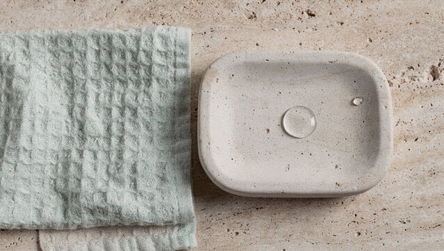 Top view of a beige ceramic soap dish with liquid drops next to a light green waffle towel on a travertine surface