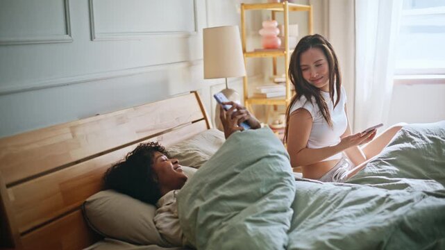 Young women relaxing bed using phones. Multiracial roommates morning bedroom.