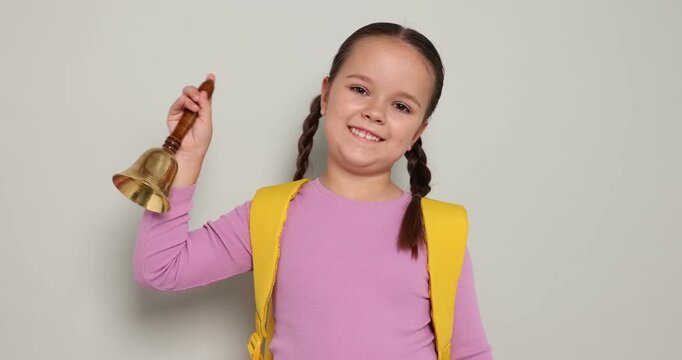 Cute girl ringing school bell on light grey background