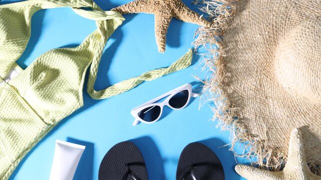 Straw hat, flip-flops, swimsuit and sunglasses on light blue background, flat lay. Camera moving around