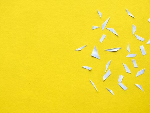Top-down minimalist view of small white geometric paper shards and clippings scattered across a solid bright yellow background.