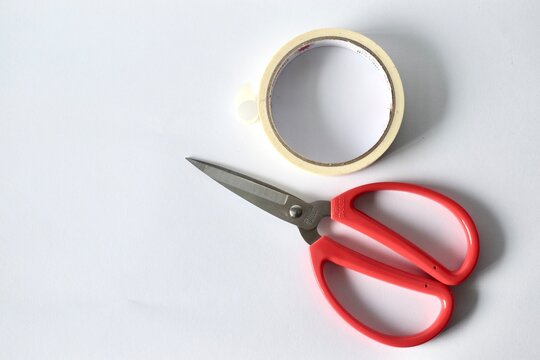 Close-up flat lay of red utility scissors and an adhesive masking tape roll arranged closely on a clean white table surface.
