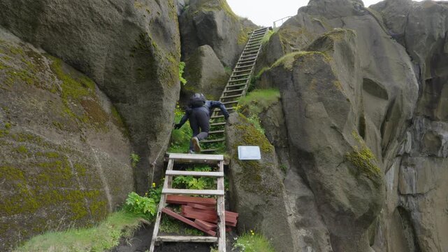 An adventurous hiker ascends a precarious wooden ladder on a steep volcanic rock trail up Heimaklettur mountain on Iceland's Vestmannaeyjar island. Ideal for travel and outdoor fitness content.