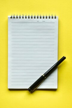 A modern black ink pen placed diagonally across the bottom corner of an open, blank lined notepad on a vivid yellow surface.