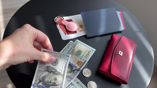 Female hands counting US dollar bills and placing them on a black round table next to a red wallet, passport, coins, and travel essentials in a horizontal 4K video.