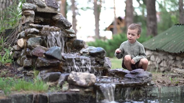 Young boy crouches by a stone waterfall in a pine forest, watching cascading water with quiet curiosity. Serene outdoor nature scene.