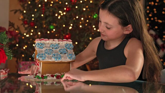A girl decorates a gingerbread house with icing and candy at a table near a Christmas tree.