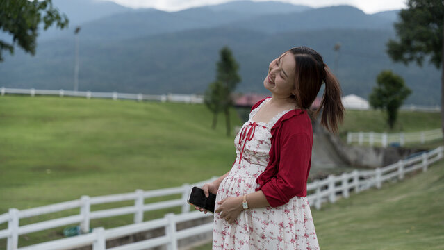 Joyful pregnant woman in a floral dress laughs with hair flowing, holding her phone and belly, on a farm with mountains behind.
