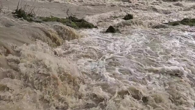 Fast flowing muddy brown flood water churning dangerously down a swollen riverbank.