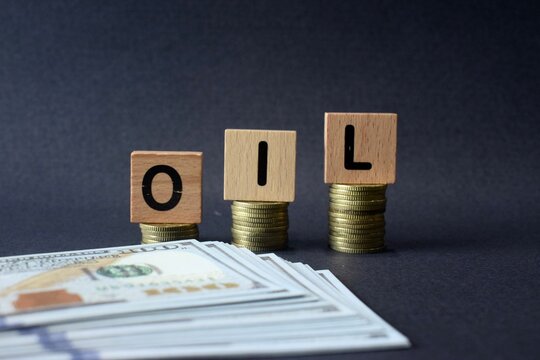 A close-up shot of the word OIL spelled out on wooden blocks atop coin stacks, with US hundred-dollar bills filling the lower foreground.