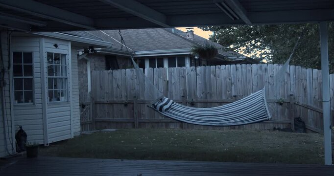 An empty hammock swings vigorously in strong wind across a fenced suburban backyard at dusk. A moody outdoor scene capturing powerful weather, movement, and quiet residential surroundings. Louisiana.