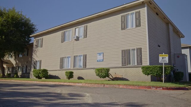 Sunny daytime view of a beige multifamily housing building with shuttered windows, security sign, and landscaped lawn.