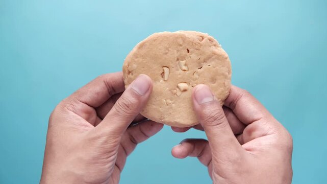 Hands break biscuit in half, revealing cashew nut pieces, close up, against blue background